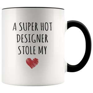 May include: White ceramic mug with a black handle and rim. The mug features the text "A SUPER HOT DESIGNER STOLE MY" in black, with a red heart graphic below the text. A humorous gift idea.