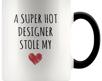 May include: White ceramic mug with a black handle and rim. The mug features the text "A SUPER HOT DESIGNER STOLE MY" in black, with a red heart graphic below the text. A humorous gift idea.