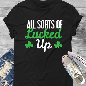 Könnte beinhalten: Schwarzes T-Shirt mit weißem Text, der "All Sorts of Lucked Up" lautet, mit grünen Kleeblatt-Symbolen auf beiden Seiten des Textes.