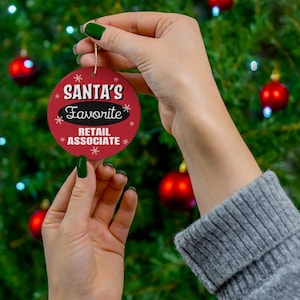 May include: Red and white Christmas ornament with the text "Santa's Favorite Retail Associate" in black lettering.