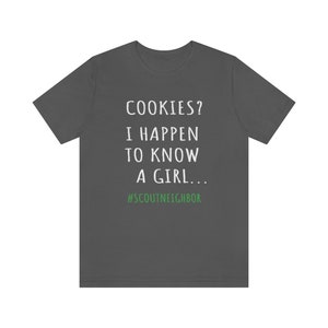 May include: A dark gray t-shirt with white text that reads "COOKIES? I HAPPEN TO KNOW A GIRL... #SCOUTNEIGHBOR".