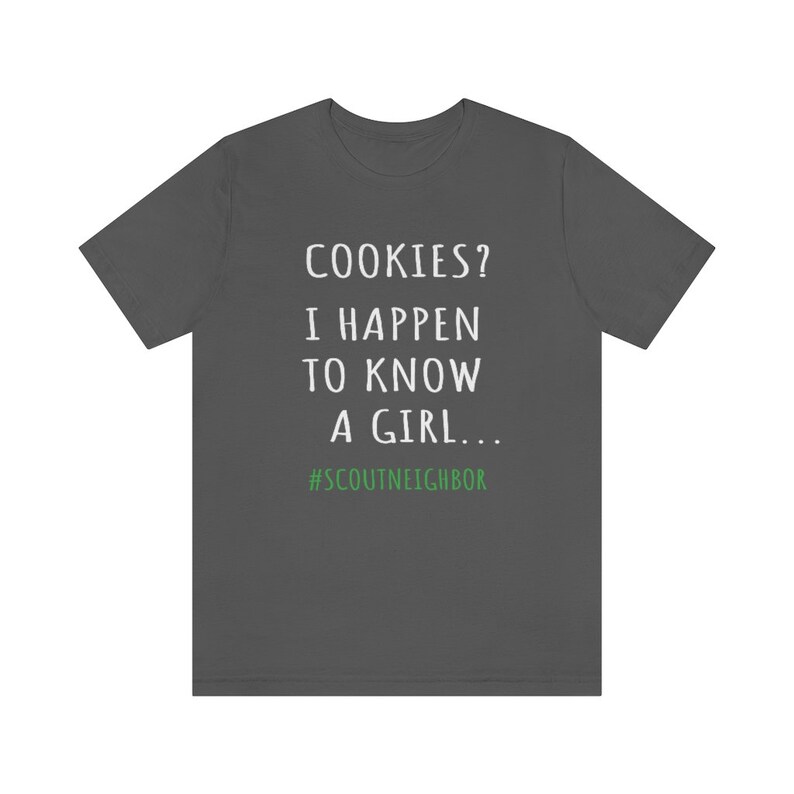 May include: A dark gray t-shirt with white text that reads "COOKIES? I HAPPEN TO KNOW A GIRL... #SCOUTNEIGHBOR".