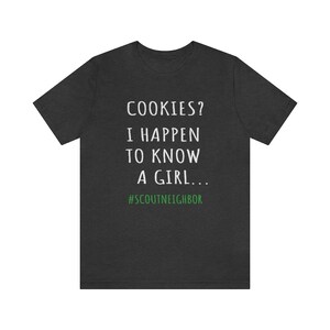 May include: Dark gray t-shirt with white text that reads "COOKIES? I HAPPEN TO KNOW A GIRL... #SCOUTNEIGHBOR".