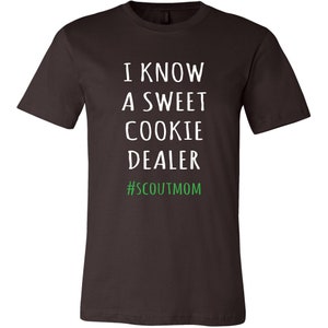 May include: A dark brown t-shirt with white text that reads "I know a sweet cookie dealer #scoutmom" in green.