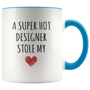 May include: White ceramic mug with a blue interior, handle, and rim. The mug features the text "A SUPER HOT DESIGNER STOLE MY" in black, with a red heart graphic below the text. A fun gift for a designer.