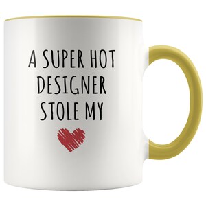 May include: White ceramic mug with a yellow handle and rim. The mug features the text "A SUPER HOT DESIGNER STOLE MY" in black, with a red heart graphic below the text. The mug is designed for hot beverages.