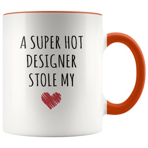 May include: White ceramic mug with an orange interior and handle. The mug features the text "A SUPER HOT DESIGNER STOLE MY" in black, with a red heart graphic below the text. A fun gift for a designer.