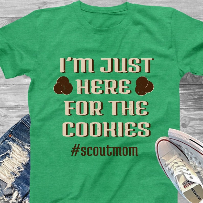 May include: Green t-shirt with the text "I'm just here for the cookies #scoutmom" in brown and beige lettering. Two brown cookie icons are on either side of the word "here".