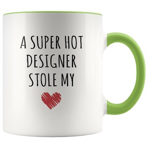 May include: White ceramic mug with a green handle and interior rim. The mug features the text "A SUPER HOT DESIGNER STOLE MY" in black, with a red heart graphic below the text. A fun gift for a designer.