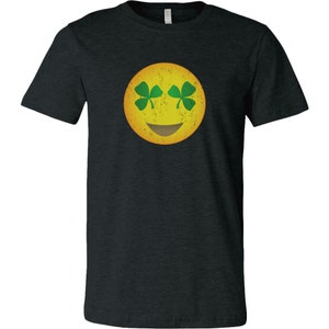Könnte beinhalten: Dunkelgraues T-Shirt mit einem gelben Emoji-Gesicht, grünen Kleeblatt-Augen und einem braunen Lächeln. Das Design hat einen Used-Look im Vintage-Stil. Ein lässiges T-Shirt für St. Patrick's Day oder den Alltag.