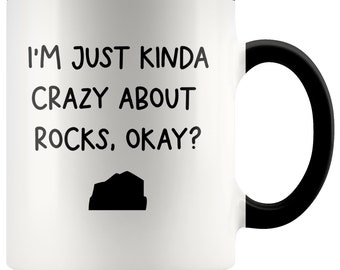 May include: White ceramic coffee mug with a black rim and handle. The mug has black text that reads "I'm just kinda crazy about rocks, okay?" with a small black silhouette of a rock below the text.