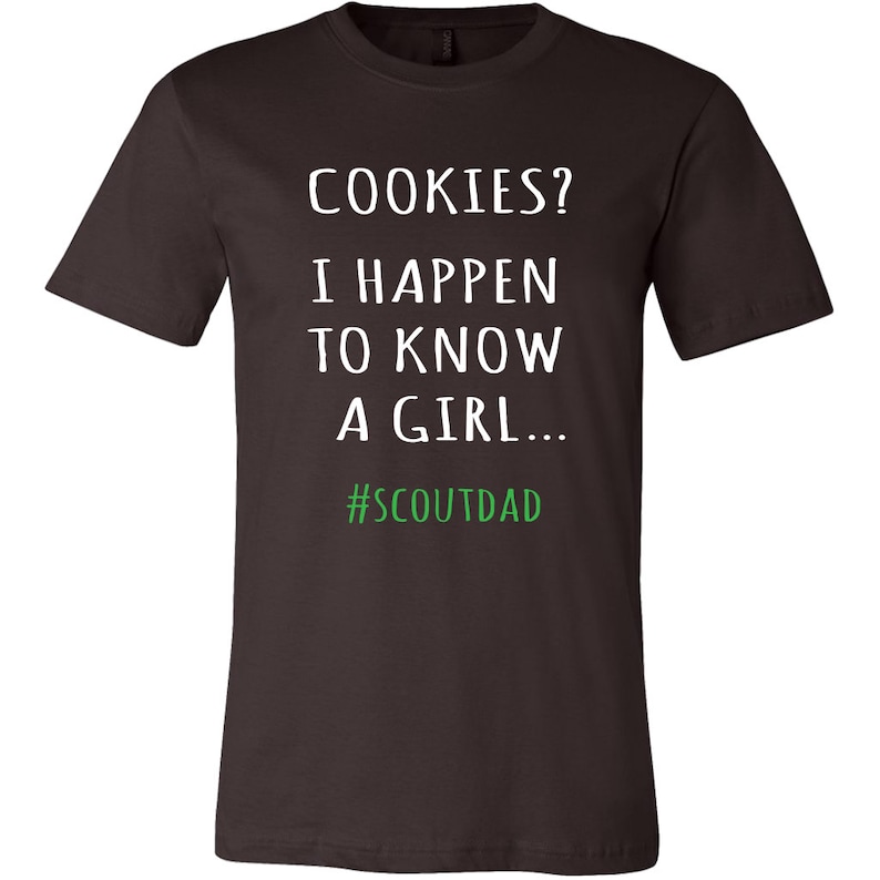 May include: Dark brown t-shirt featuring the text "COOKIES? I HAPPEN TO KNOW A GIRL..." in white, with the hashtag "#SCOUTDAD" in green. A casual wear item.