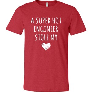 May include: Red t-shirt with white text that reads "A SUPER HOT ENGINEER STOLE MY" above a white heart graphic. The shirt is a solid red color and has short sleeves.
