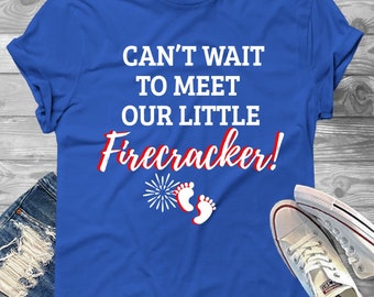 May include: A blue t-shirt with the text "Can't wait to meet our little Firecracker!" in white with red accents. The shirt features a red, white, and blue firework graphic and a pair of white baby footprints.