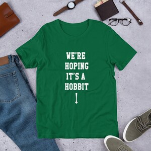 Może przedstawiać: Zielony T-shirt z białym tekstem, kt&oacute;ry brzmi "We're Hoping It's a Hobbit" ze strzałką skierowaną w d&oacute;ł.