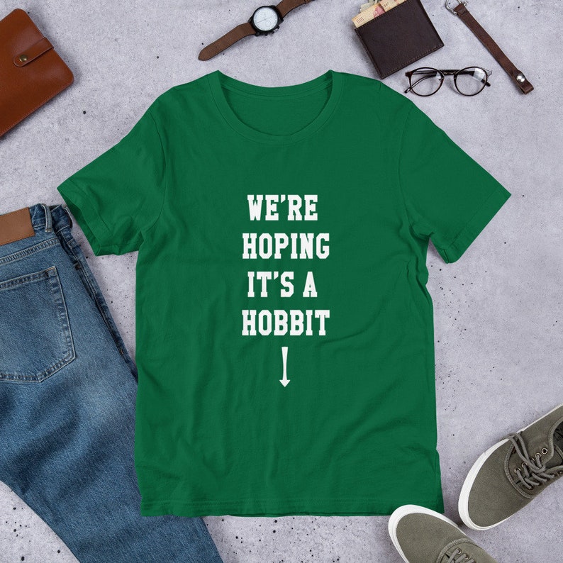Może przedstawiać: Zielony T-shirt z białym tekstem, kt&oacute;ry brzmi "We're Hoping It's a Hobbit" ze strzałką skierowaną w d&oacute;ł.