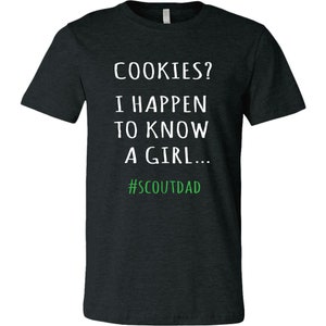 May include: Dark grey t-shirt with white text saying "COOKIES? I HAPPEN TO KNOW A GIRL..." and the hashtag "#SCOUTDAD" in green. The shirt has a crew neck.