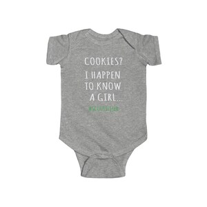 May include: Gray baby onesie with the text "COOKIES? I HAPPEN TO KNOW A GIRL... #SCOUTSISISTER" in white. The text "#SCOUTSISISTER" is in green.