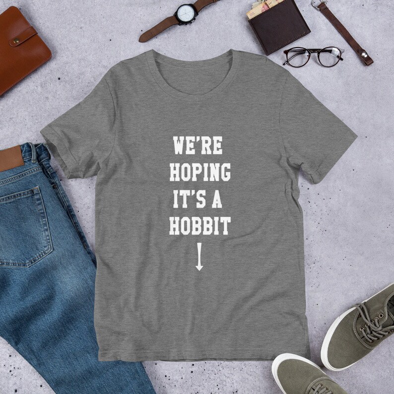 Może przedstawiać: Szary t-shirt z nadrukiem "We're Hoping It's A Hobbit" i strzałką skierowaną w d&oacute;ł, w kolorze białym.