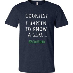 May include: A navy blue t-shirt featuring the white text: "COOKIES? I HAPPEN TO KNOW A GIRL..." with the green hashtag "#SCOUTDAD". The shirt has a crew neck.