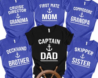 May include: A collection of blue and black t-shirts with nautical-themed text. The shirts feature phrases like "Cruise Director Grandma," "First Mate Mom," and "Captain Dad," along with anchor symbols. A wooden ship's wheel is in the foreground.