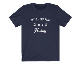 May include: Navy blue t-shirt with white text that reads "My therapist is a Husky" with two white paw prints on either side of the word "is".