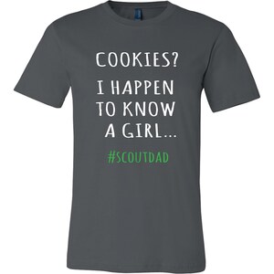 May include: A dark grey t-shirt featuring the text "COOKIES? I HAPPEN TO KNOW A GIRL..." in white, with the hashtag "#SCOUTDAD" in green. The shirt has a crew neck.