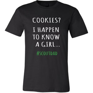 May include: Black t-shirt featuring the text "COOKIES? I HAPPEN TO KNOW A GIRL..." in white, with the hashtag "#SCOUTDAD" in green. The shirt has a crew neck.