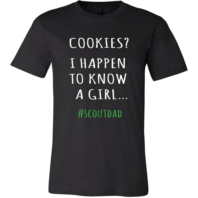 May include: Black t-shirt featuring the text "COOKIES? I HAPPEN TO KNOW A GIRL..." in white, with the hashtag "#SCOUTDAD" in green. The shirt has a crew neck.