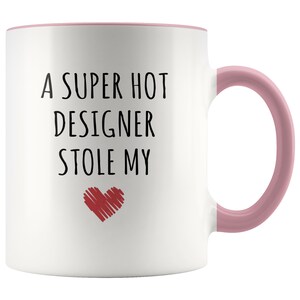 May include: White ceramic mug with a pink interior and handle. The mug features the text "A SUPER HOT DESIGNER STOLE MY" in black, with a red heart graphic below the text. A fun gift for a designer.