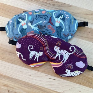 May include: Two sleep masks, one with a purple background and a pattern of white cats and rainbows, and the other with a blue background and a pattern of mermaids and sea life.
