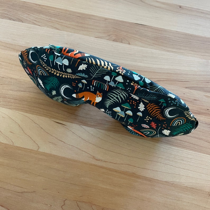 May include: A dark-colored, fabric-covered eye mask featuring a woodland pattern. The pattern includes foxes, mushrooms, ferns, rainbows, and crescent moons in shades of orange, green, white, and gold. The mask is designed to block out light for sleeping or relaxation.