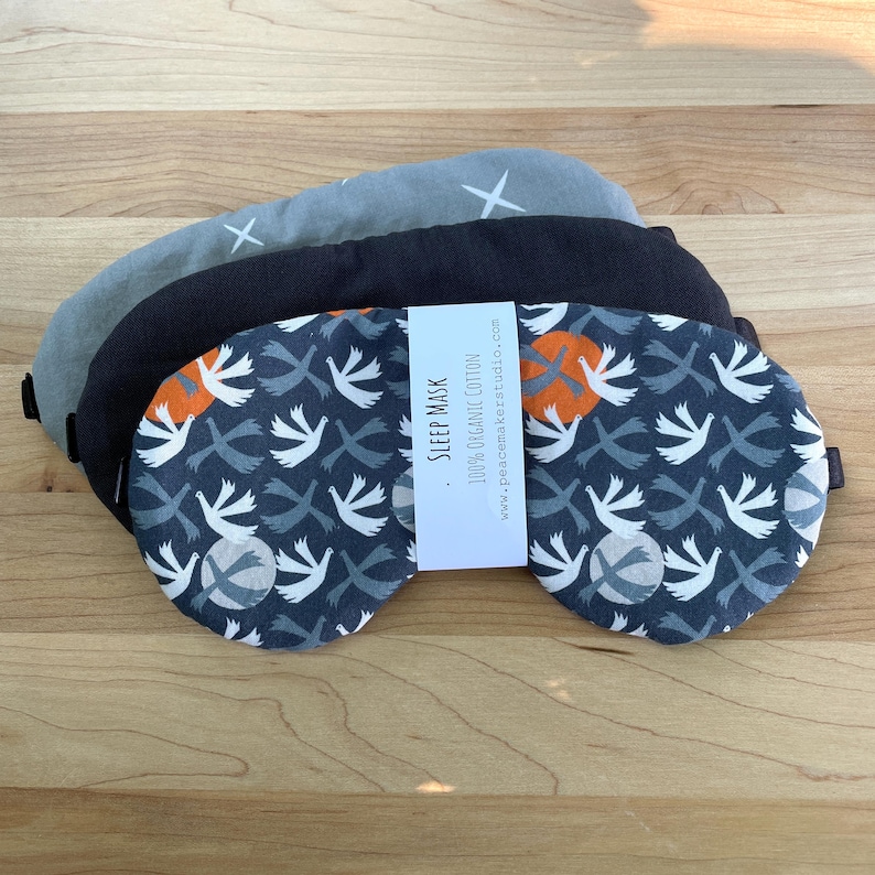 May include: Three sleep masks in gray, black, and patterned navy blue. The navy mask features a white bird and orange circle design. The gray mask has white cross accents. A white label reads "Sleep Mask" and "100% Organic Cotton."