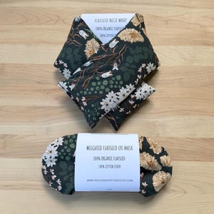 Pode incluir: Duas compressas de sementes de linho verde escuro com estampas florais. Uma é uma compressa para o pescoço, a outra é uma máscara para os olhos. A compressa para o pescoço é dobrada e a máscara para os olhos tem formato oval. Ambas têm etiquetas brancas com informações do produto.