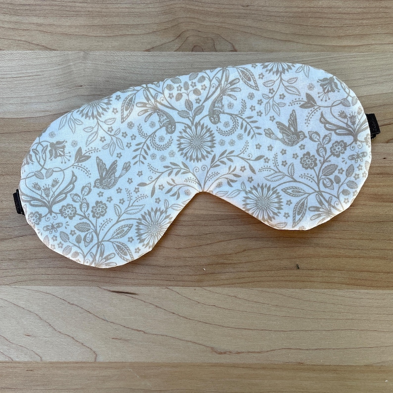 May include: A white sleep mask with a floral and bird pattern in shades of tan and gray. The mask has a black elastic strap and is designed to cover the eyes for light blocking.