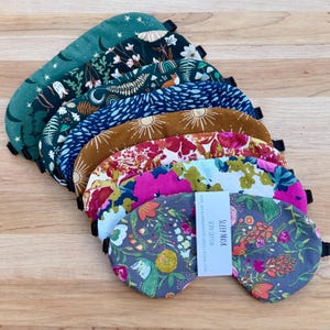 May include: A stack of eight fabric sleep masks featuring various patterns including celestial motifs, floral prints, and woodland themes. Each mask has a black elastic strap. A white tag on the top mask reads: SLEEP MASK 100% COTTON peacemakerstudio.etsy.com.