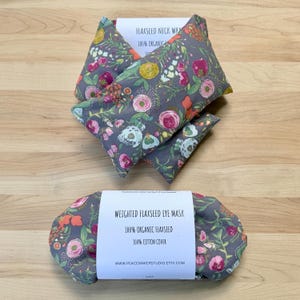 Pode incluir: Envoltórios para o pescoço e máscara para os olhos de linho com estampa floral cinza. Os envoltórios para o pescoço são triangulares e a máscara para os olhos é oval. Os produtos são rotulados com o texto "100% Organic Flaxseed" e "100% Cotton Cover".