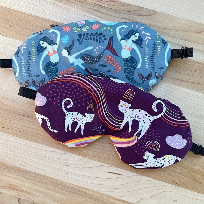 May include: Two sleep masks, one with a purple background and a pattern of white cats and rainbows, and the other with a blue background and a pattern of mermaids and sea life.