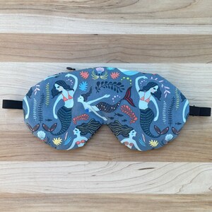 May include: A blue sleep mask with a mermaid pattern. The mask features three mermaids with long flowing hair and tails, surrounded by seaweed and other ocean-themed details.