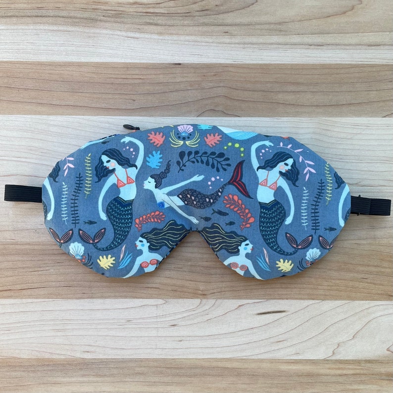 May include: A blue sleep mask with a mermaid pattern. The mask features three mermaids with long flowing hair and tails, surrounded by seaweed and other ocean-themed details.