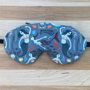 Puede incluir: Una m&aacute;scara para dormir azul con un estampado de sirenas. La m&aacute;scara presenta sirenas con cabello negro y tops rojos, rodeadas de plantas marinas y criaturas marinas. La m&aacute;scara tiene una banda el&aacute;stica negra.