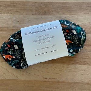 May include: A dark eye mask with a whimsical forest pattern featuring foxes, rainbows, and mushrooms. The mask is wrapped with a white label that reads "WEIGHTED FLAXSEED & LAVENDER EYE MASK". The mask is made of 75% organic flaxseed, 25% dried organic lavender flowers, and a 100% cotton cover.