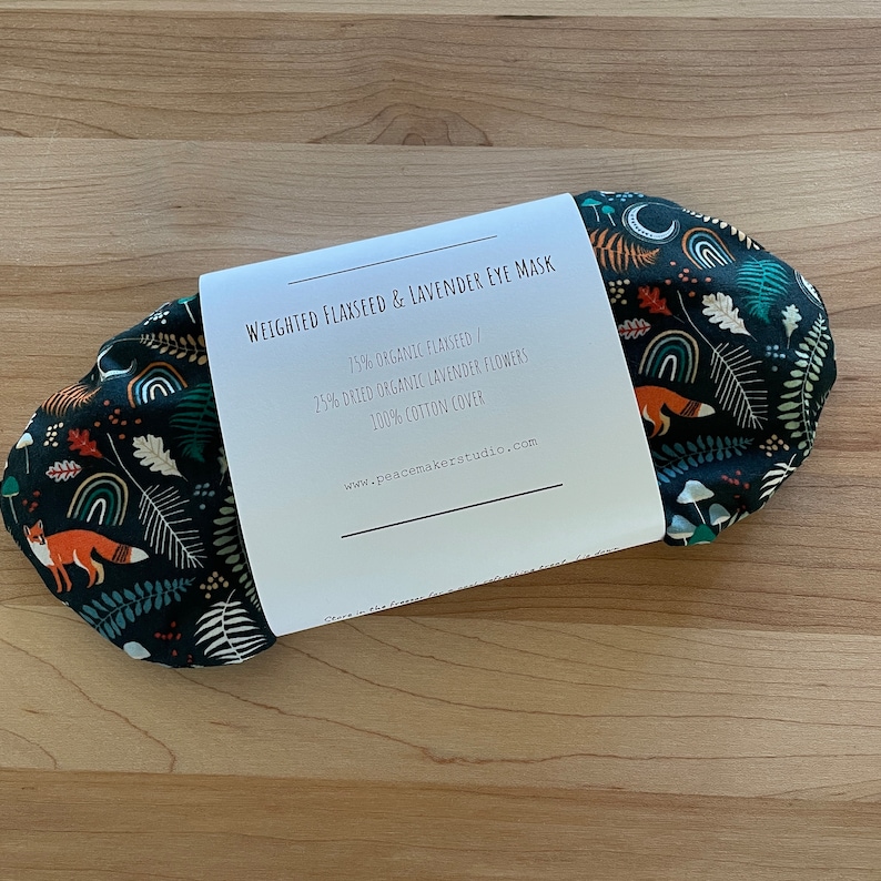May include: A dark eye mask with a whimsical forest pattern featuring foxes, rainbows, and mushrooms. The mask is wrapped with a white label that reads "WEIGHTED FLAXSEED & LAVENDER EYE MASK". The mask is made of 75% organic flaxseed, 25% dried organic lavender flowers, and a 100% cotton cover.