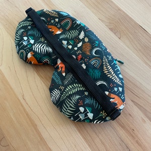 May include: A dark green sleep mask with a woodland pattern featuring orange foxes, rainbows, mushrooms, and ferns. The mask has a black elastic strap and a zipper. The mask is designed to block out light.