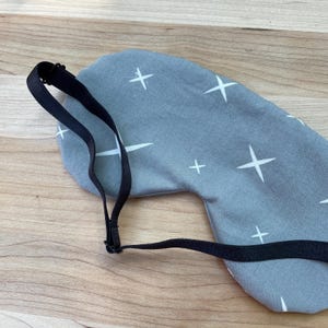 May include: A gray sleep mask with a white star pattern and a black adjustable strap. The mask is designed to block out light for better sleep. The background is a light wood surface.