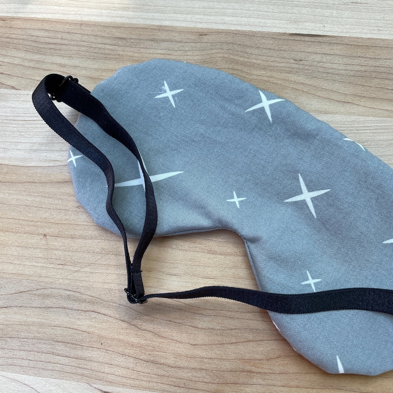 May include: A gray sleep mask with a white star pattern and a black adjustable strap. The mask is designed to block out light for better sleep. The background is a light wood surface.