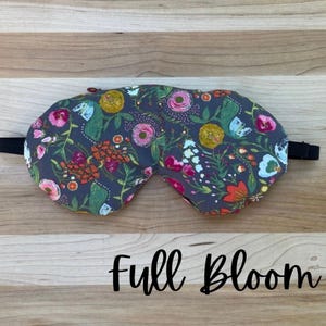 Puede incluir: Una m&aacute;scara para dormir floral con un fondo gris oscuro y una correa el&aacute;stica negra. La m&aacute;scara presenta un patr&oacute;n colorido de flores en tonos de rosa, naranja, amarillo y azul. El texto "Full Bloom" est&aacute; escrito en cursiva.