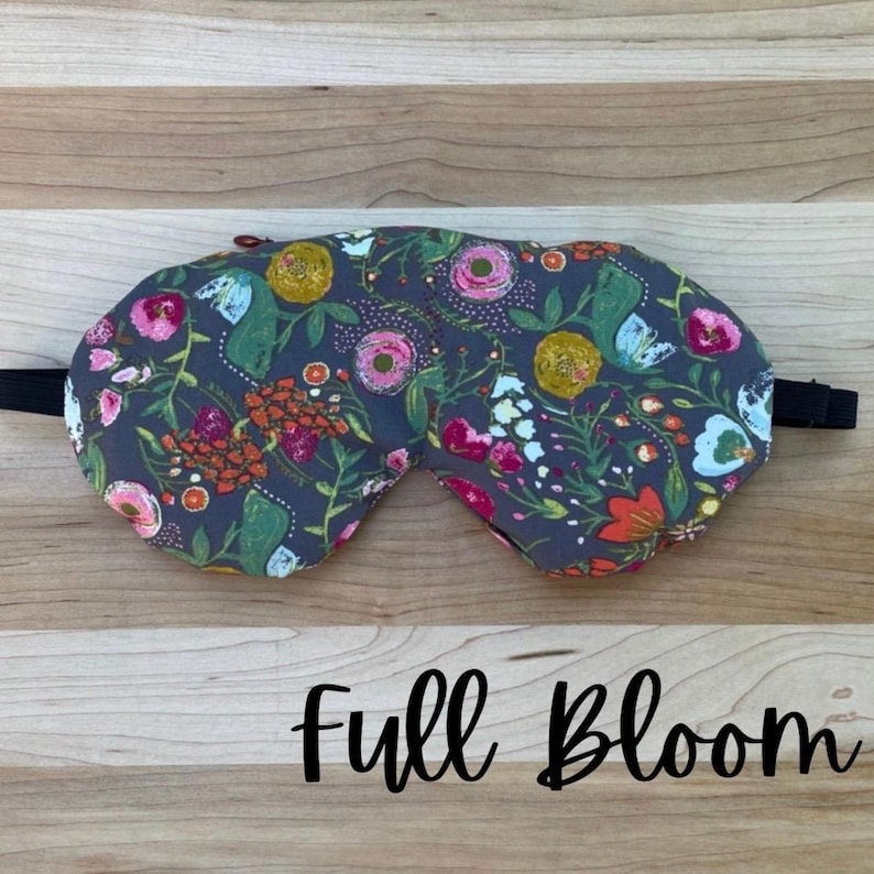 Puede incluir: Una m&aacute;scara para dormir floral con un fondo gris oscuro y una correa el&aacute;stica negra. La m&aacute;scara presenta un patr&oacute;n colorido de flores en tonos de rosa, naranja, amarillo y azul. El texto "Full Bloom" est&aacute; escrito en cursiva.