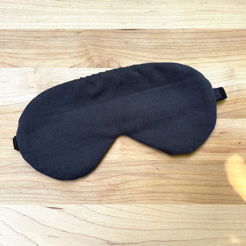 May include: A dark gray sleep mask with a curved design. The mask has a strap on the side for securing around the head. The mask is made of a soft, textured fabric. The sleep mask is designed to block out light.