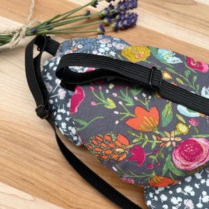 May include: A black sleep mask with an adjustable strap. The mask is made of fabric with a floral pattern in shades of pink, orange, yellow, and green on a dark gray background.